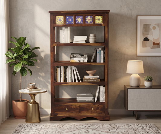 A tall, dark brown wooden bookshelf featuring a row of decorative blue and yellow patterned tiles along the top trim. It has four open shelves styled with books and decorative bowls, with two small storage drawers featuring round knobs at the base. The bookcase stands in a well-lit living room setting next to a potted ficus plant and a side table.